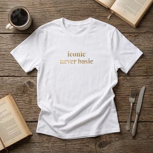 Op de afbeelding: Een wit t-shirt met korte mouwen en de tekst "iconic never basic" in een metallic gouden lettertype op het midden van de borst. Het kledingstuk heeft een ronde hals en een standaard pasvorm, plat neergelegd op een houten ondergrond.