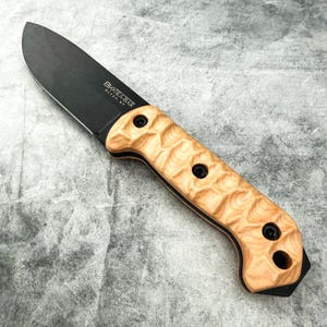Könnte beinhalten: Ein Messer mit schwarzer Klinge und hellem Holzgriff. Auf der Klinge steht "BK&T KA-BAR OLEAN, NY". Der Griff hat drei schwarze Nieten und ein geschwungenes Design.