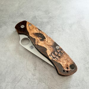 May include: A folding pocket knife with a stainless steel blade and a handle featuring a carved bear design. The handle is brown with a lighter brown bear carving. The knife has a round hole near the blade for one-handed opening.