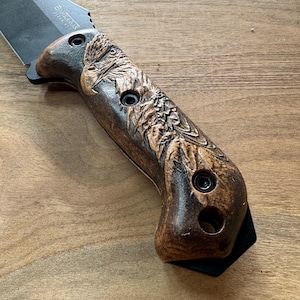 Könnte beinhalten: Nahaufnahme eines schwarzen Messers mit einem dunkelbraunen Griff. Der Griff ist mit einem Adler-Design versehen. Die Messerklinge ist schwarz und trägt den Text "BUSHBAR".