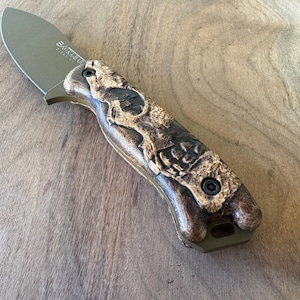 May include: A brown and black folding knife with a skull design on the handle. The knife has a black blade and a brown handle with a skull design. The knife is closed and the blade is not visible.