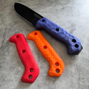 May include: A black fixed-blade knife with a purple handle and two additional replacement handles in red and orange. The knife blade has the text "BK7 EXTRA-BAR" printed on it. The handles have a textured design with holes for attachment.
