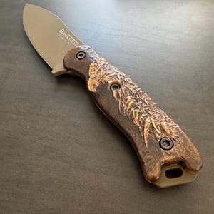 May include: A brown and black pocket knife with a wood handle featuring a carved eagle design. The blade is made of stainless steel and has the text "Black Bear" and "Outdoor Gear" engraved on it.