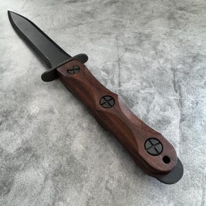 Puede incluir: Un cuchillo de hoja oscura con mango de madera. La hoja es negra y el mango es de un marrón intenso con detalles circulares negros. El cuchillo tiene guarda y un extremo redondeado. El diseño es simétrico y parece bien elaborado.