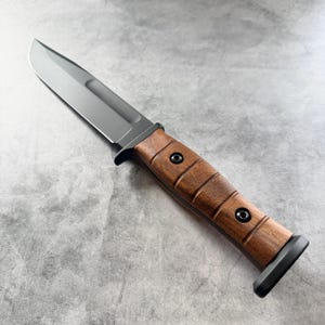 May include: A tactical knife with a dark gray blade and a brown wooden handle. The handle has a series of grooves and is secured with black rivets. The knife has a black bolster and a full tang construction.