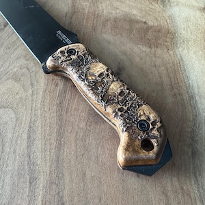 Op de afbeelding: Een zwart mes met een houten handvat met een schedelontwerp. Het handvat is gegraveerd met het woord "Satterlee" en de tekst "Blade #7".