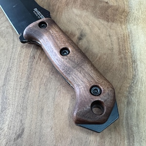 May include: Close-up of a brown wooden knife handle with black screws and a black metal ring. The handle is attached to a black knife blade.