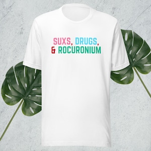 Może przedstawiać: Biała koszulka z nadrukiem "SUXS, DRUGS, & ROCURONIUM" w kolorze różowym, niebieskim i zielonym.