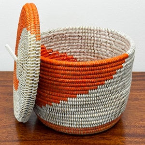 May include: A round woven basket with a matching lid, featuring a coiled construction in bright orange and off-white fibers. The basket has a deep cylindrical body and a slightly domed lid with a small white loop handle attached to the side.