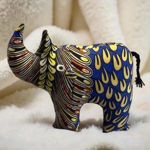 May include: A stuffed rhino toy with a vibrant patterned fabric in blue, yellow, orange, and black. The rhino has a black braided tail and a white eye. The fabric design features a mix of abstract shapes and patterns, creating a unique and colorful appearance.