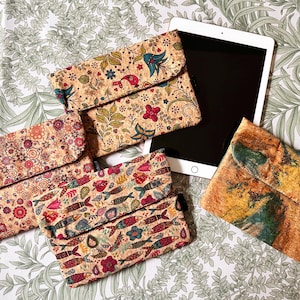 May include: A selection of cork tablet cases featuring vibrant floral, bird, and fish designs. A white tablet is placed beside the cases on a patterned fabric. The cases have a flap closure.