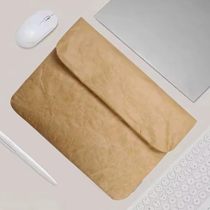Puede incluir: Una funda para portátil de color beige con cierre de solapa, un ratón inalámbrico blanco, un bolígrafo blanco, un portátil plateado y un teclado blanco están dispuestos sobre una superficie gris claro. La funda tiene un aspecto texturizado.