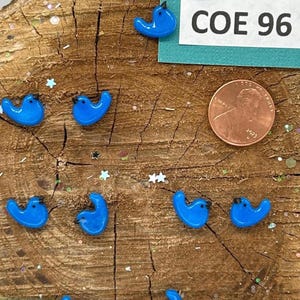 May include: A collection of small, glossy blue bird-shaped beads with black beaks. The beads are arranged on a wooden surface with a penny and a ruler for size comparison. The text "COE 96" is visible on a green card.