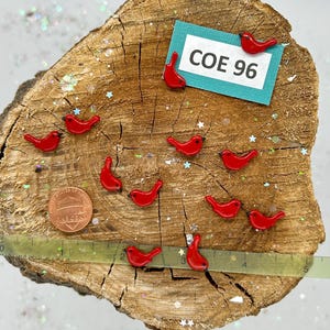 May include: A collection of 10 red glass bird figurines on a wooden surface. The birds are approximately 1 inch in size. A ruler is visible in the bottom of the image. A sign reads "COE 96".