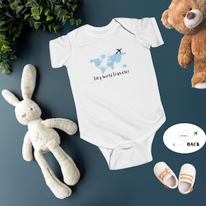 May include: White baby onesie with a blue world map design, a heart-shaped dotted line, and a black airplane graphic. The text "Tiny World Traveler" is printed below the map. A stuffed bunny, teddy bear, and baby shoes are also in the image.