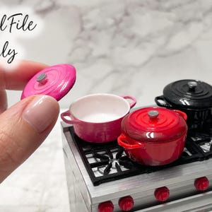 Puede incluir: Juego de cocina en miniatura con una estufa y varias ollas coloridas. El juego incluye una tapa de olla rosa, una olla rosa, una olla roja y una olla negra. La estufa tiene un acabado plateado con perillas rojas y una encimera negra.