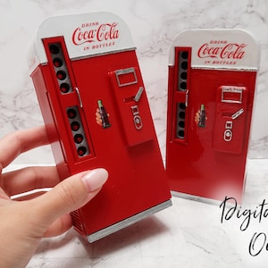 May include: Two red and white Coca-Cola vending machines with a silver trim. The vending machines are miniature and have a glass front with a Coca-Cola bottle graphic. The text "Drink Coca-Cola in Bottles" is printed on the front of the vending machines.