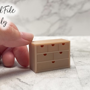 May include: A miniature beige chest of drawers with six drawers, each with a small, red-orange accent. The text "Digital File Only" is in the upper left corner. The chest of drawers is held by a hand.