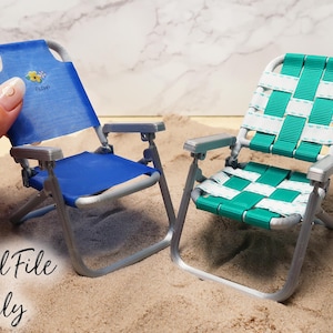 May include: Two miniature folding beach chairs, one blue and one green, on a sandy surface. The blue chair has a floral design on the back.