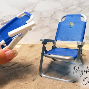 May include: Two miniature blue and white beach chairs. One chair is folded and the other is unfolded. The chairs are sitting on a sandy surface.