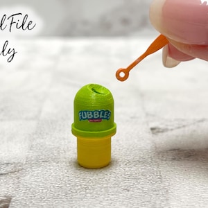 Op de afbeelding: Een groene en gele plastic zeepbelstok met het woord "FUBBLES" erop gedrukt. De stok wordt vastgehouden door een hand met een vinger die naar de stok wijst.