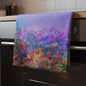 Puede incluir: Un paño de cocina con un diseño floral. El paño presenta una pintura impresionista vibrante de flores silvestres coloridas en tonos de rojo, rosa, amarillo y azul sobre un fondo lavanda. El paño está colgado de un asa de horno.