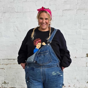 Peut inclure: Une personne portant un sweat-shirt noir avec un dessin d'une femme fléchissant son biceps, un bandana rose et une salopette en jean bleu. La salopette a des éclaboussures de peinture. Le fond est un mur de briques blanches.