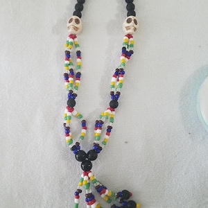 Op de afbeelding: Een kralenketting met een zwarte lavasteen ketting en kleurrijke kralen. De ketting heeft twee schedelvormige hangers en een kwast van zwarte kralen. De kralen zijn in verschillende kleuren, waaronder rood, geel, blauw en groen.