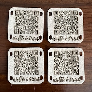 May include: Four white square wooden tags with black and white QR codes and the text "Waffle & Stitch" printed on each tag.