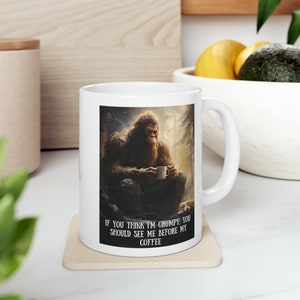 May include: White ceramic mug with a brown Bigfoot illustration holding a cup of coffee. The mug has the text "If you think I'm grumpy, you should see me before my coffee" printed on the bottom.