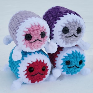 Peut inclure: Quatre créatures faites à la main au crochet, dans les tons de bleu, rouge, violet et rose. Chacune a une bordure blanche, des yeux noirs et une bouche souriante. Les créatures sont empilées par paires, deux sur deux.