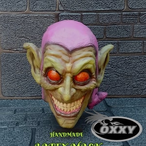 May include: Green Goblin latex mask with a green face, red eyes, and a wide, toothy grin. The mask is handmade and has a pink hood. The mask is displayed against a brick wall with the text "GREEN GOBLIN FANART" and "HANDMADE LATEX MASK" below it.