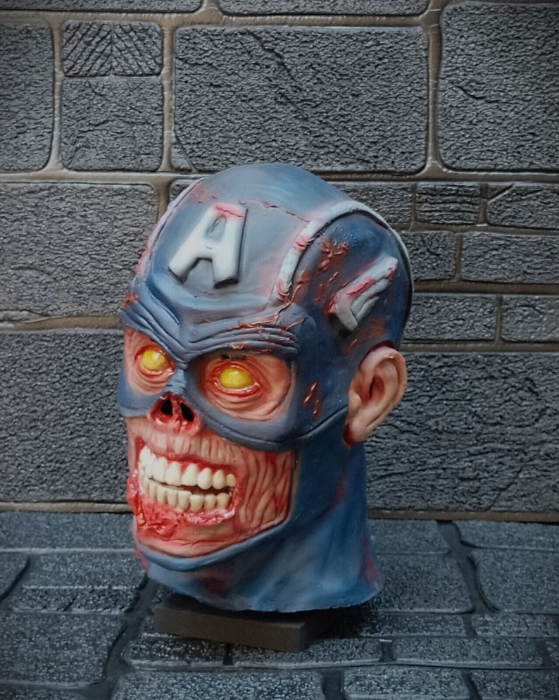 May include: A blue and red zombie Captain America mask with a cracked and weathered appearance. The mask has a large, toothy grin and yellow eyes.