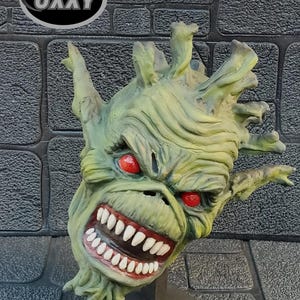 May include: A green and brown monster mask with red eyes and sharp teeth. The mask is made of a textured material and is mounted on a black stand.
