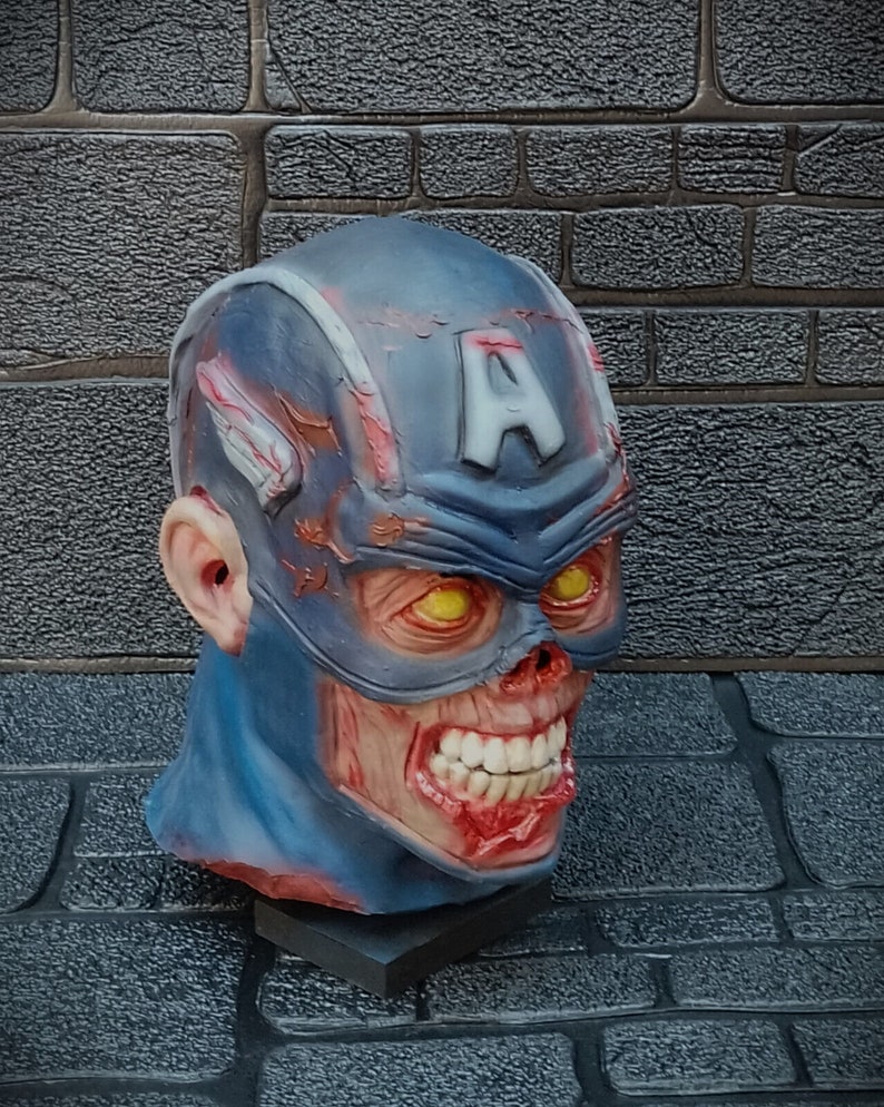 May include: A blue and red zombie Captain America mask with a cracked and weathered appearance. The mask has a large 'A' on the forehead and yellow eyes. The mouth is open and shows teeth.