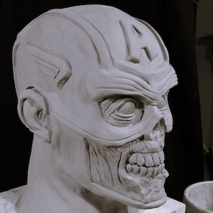 May include: A white clay sculpture of a zombie superhero head with a sculpted 'A' on the forehead. The sculpture is on a white base.