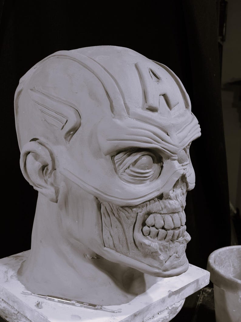 May include: A white clay sculpture of a zombie superhero head with a sculpted 'A' on the forehead. The sculpture is on a white base.
