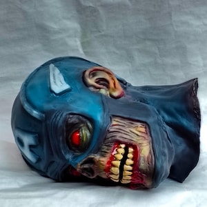 May include: A blue and white zombie Captain America mask with a red eye and a bloody mouth. The mask is made of plastic and is lying on a white surface.