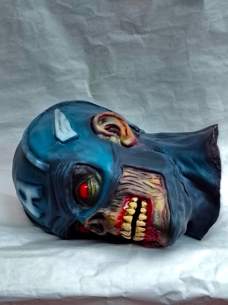 May include: A blue and white zombie Captain America mask with a red eye and a bloody mouth. The mask is made of plastic and is lying on a white surface.