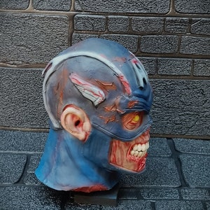 May include: A blue and red zombie Captain America mask with a cracked and weathered look. The mask has a yellow eye and a cracked, open mouth with visible teeth.