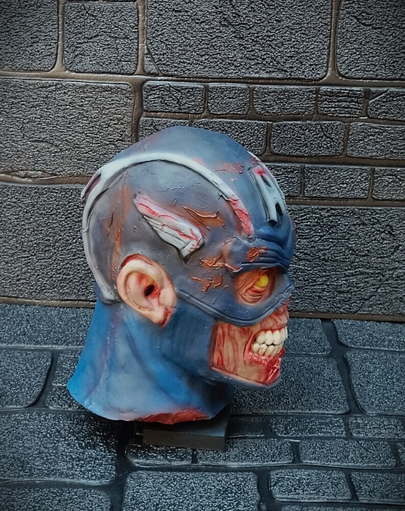 May include: A blue and red zombie Captain America mask with a cracked and weathered look. The mask has a yellow eye and a cracked, open mouth with visible teeth.