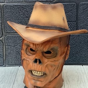 Puede incluir: Una máscara de Halloween marrón y naranja con un sombrero de vaquero. La máscara tiene una cara de calavera con una amplia sonrisa y una barba marrón.