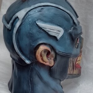 May include: A blue and white zombie Captain America Halloween mask. The mask is made of latex and has a detailed design. The mask has a distressed look with visible cracks and tears.