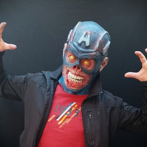 May include: A person wearing a blue and red zombie Captain America mask with glowing yellow eyes. The mask has a white letter "A" on the forehead. The person is wearing a red t-shirt with a white and yellow stripe design and a black jacket.