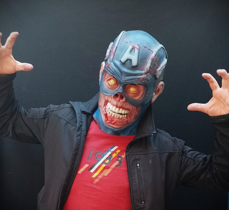 May include: A person wearing a blue and red zombie Captain America mask with glowing yellow eyes. The mask has a white letter "A" on the forehead. The person is wearing a red t-shirt with a white and yellow stripe design and a black jacket.