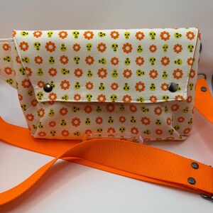 May include: A rectangular crossbody bag featuring an off-white fabric base with a repeating pattern of orange flowers and small lime green skulls. The bag includes a wide, bright orange adjustable strap and two dark metal snap button closures.