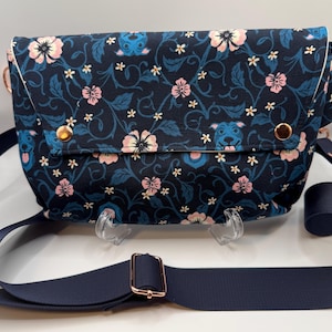 May include: A navy blue crossbody bag featuring a floral pattern with pink flowers and small blue dog faces. The bag has a fold-over flap secured with two gold-toned metal snaps and includes an adjustable navy blue fabric strap with gold hardware.