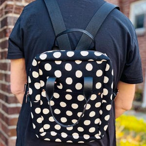 May include: Black backpack with a white polka dot pattern. The backpack has black straps, a front pocket, and black leather handles. The bag is worn on the back.