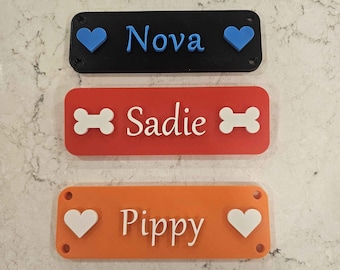 May include: Three personalized dog name tags. The top tag is black with blue hearts and the name "Nova". The middle tag is red with white bone icons and the name "Sadie". The bottom tag is orange with white hearts and the name "Pippy".