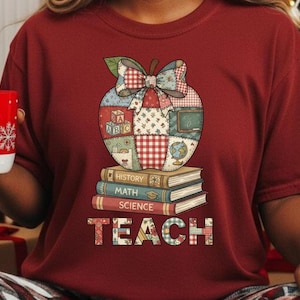 May include: Burgundy t-shirt featuring a patchwork apple design with a bow, atop a stack of books labeled "History," "Math," and "Science." The word "TEACH" is displayed in a patchwork style at the bottom. The apple design includes elements like an ABC block, a globe, and a chalkboard.